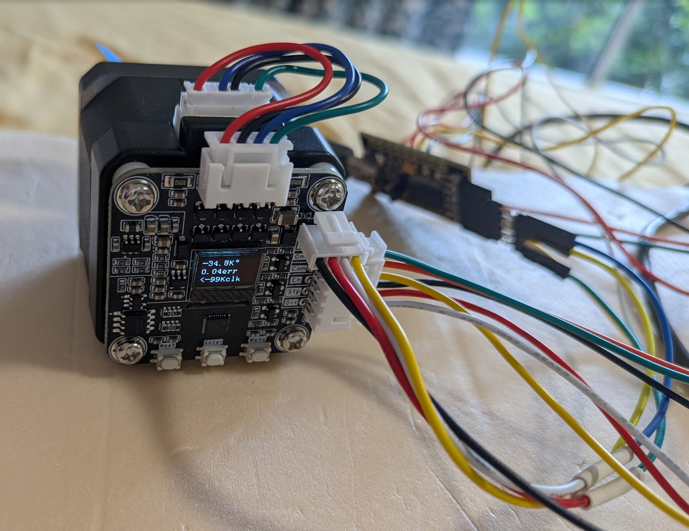 A photo of a stepper motor with various colourful wires attached. A small OLED screen on the back of the motor reads "-34.8k° 0.04err <-99Kclk". Some trees are visible through a window in the background. A photo of a stepper motor with various colourful wires attached. A small OLED screen on the back of the motor reads "-34.8k° 0.04err <-99Kclk". Some trees are visible through a window in the background.
