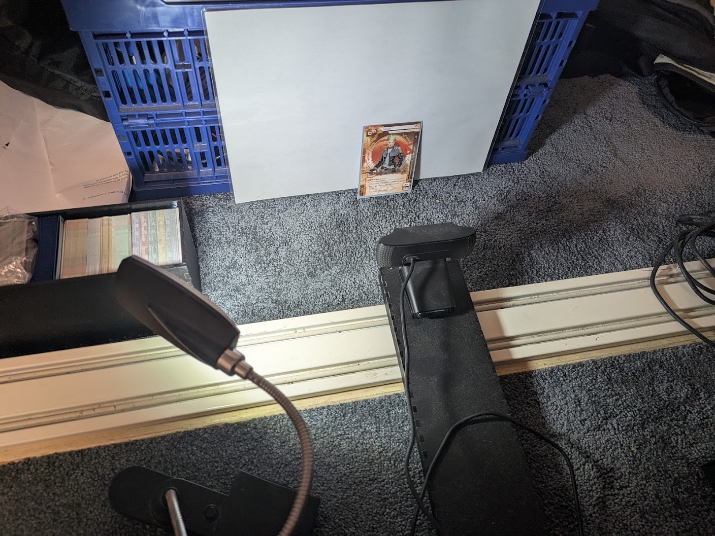 A photo of a netrunner card standing upright against a white background with carpet beneath it. A light is shining at the card from an oblique angle, and a webcam is sitting on a piece of black plastic pointing at the card. The webcam is about 150mm from the card. Other junk is visible in the background. A photo of a netrunner card standing upright against a white background with carpet beneath it. A light is shining at the card from an oblique angle, and a webcam is sitting on a piece of black plastic pointing at the card. The webcam is about 150mm from the card. Other junk is visible in the background.