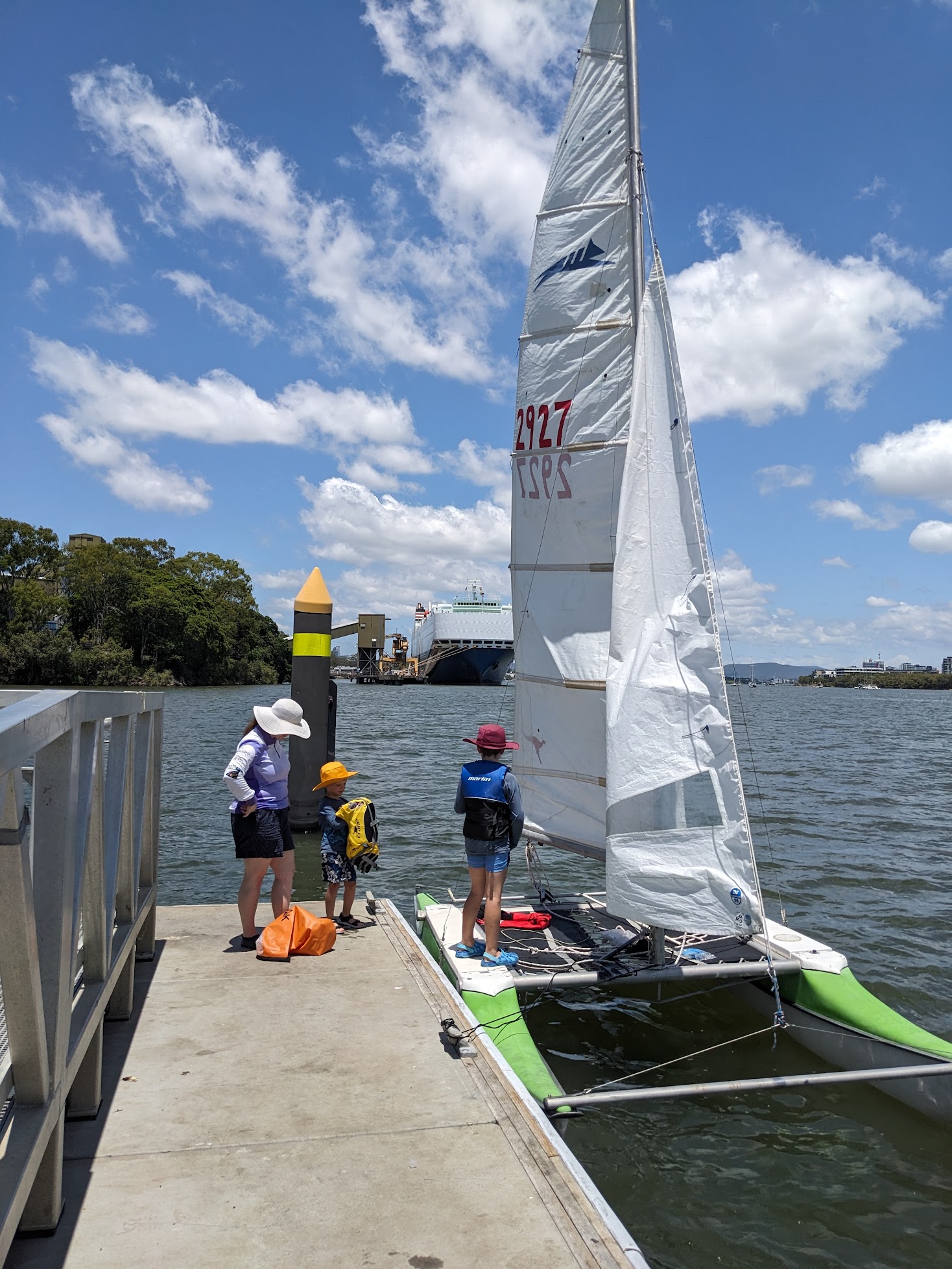 A photo of Michelle, Alex and Sam standing around the boat tied up at a dock. A large car carrier is visible in the background. The sky is littered with fluffy white clouds.