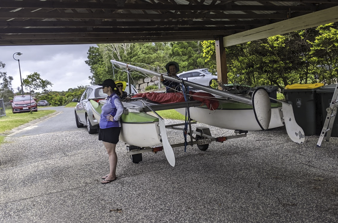 A photo of the boat on the trailer behind our silver corolla under our carport. Sam is sitting on the boat, Michelle is standing to the left and Alex is visible in the background.