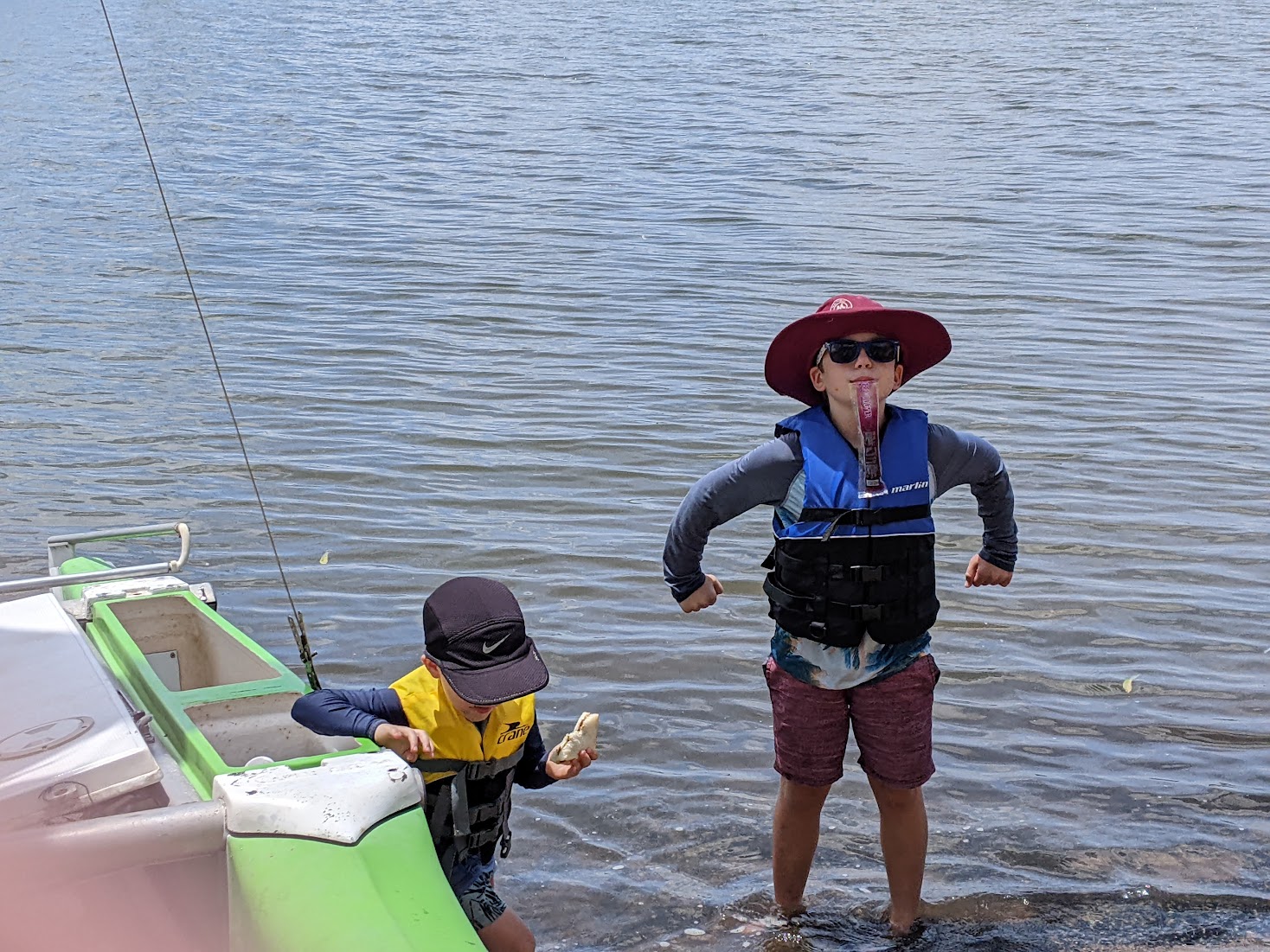 A photo of two boys standing in water next to the boat. Alex is striking a strongman pose with a zooper dooper. Sam is holding a vegemite sandwich.